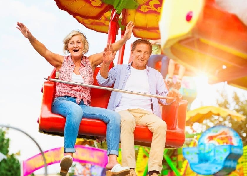 An older couple joyfully rides a colorful amusement park attraction with their arms raised. An older couple joyfully rides a colorful amusement park attraction with their arms raised.