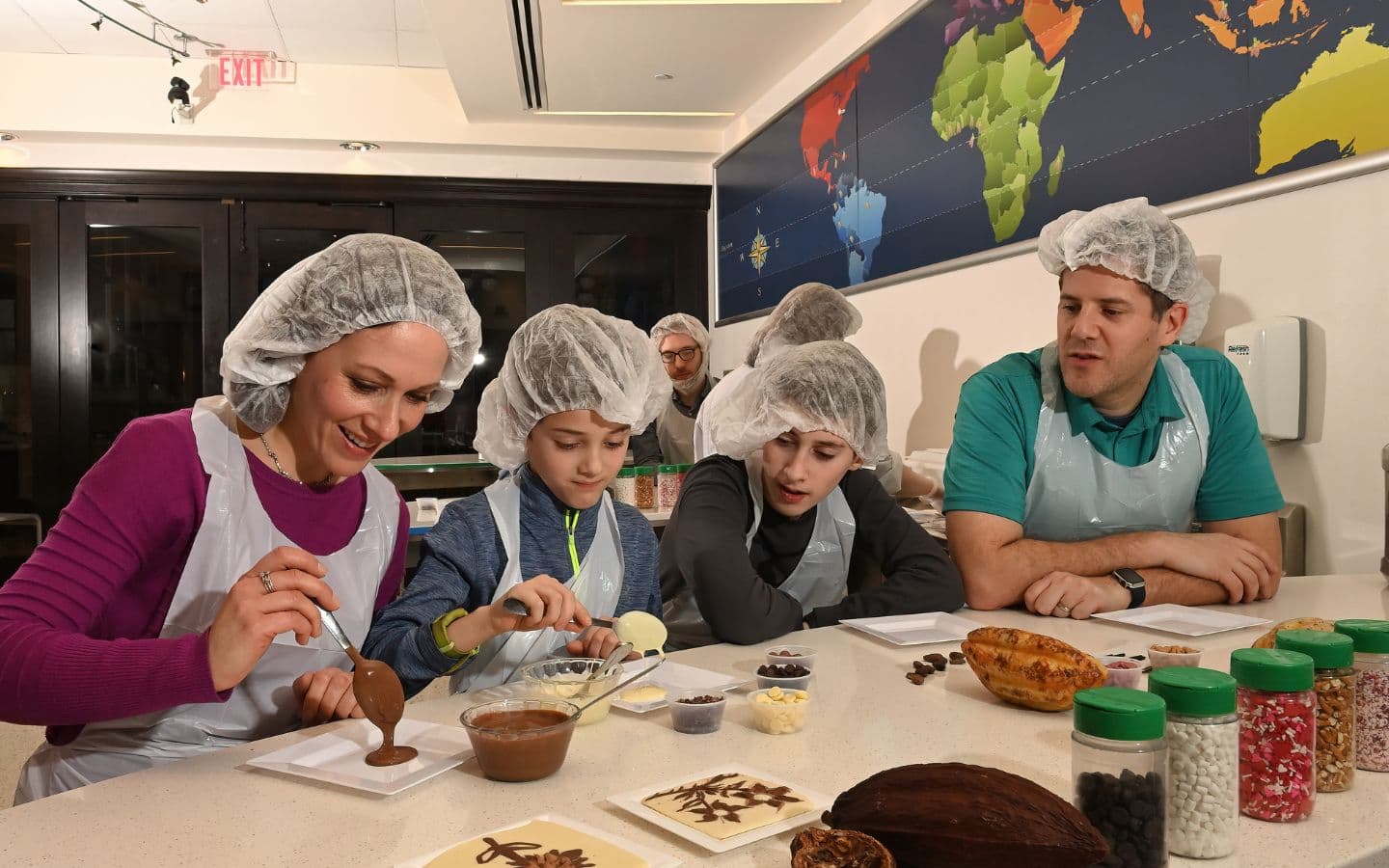 A group of four, wearing hair nets and aprons, are engaged in a chocolate-making activity at a table filled with various ingredients.