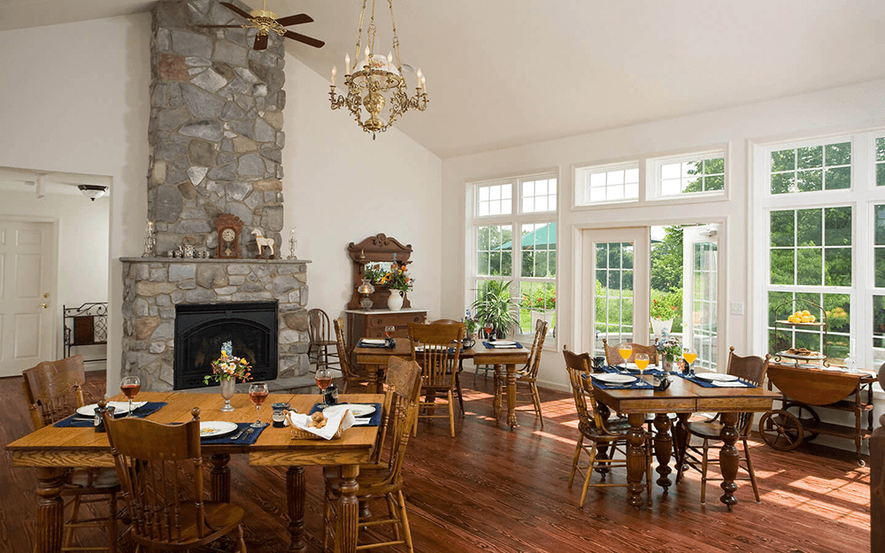 A cozy dining room featuring wooden tables, a stone fireplace, and large windows overlooking a garden.