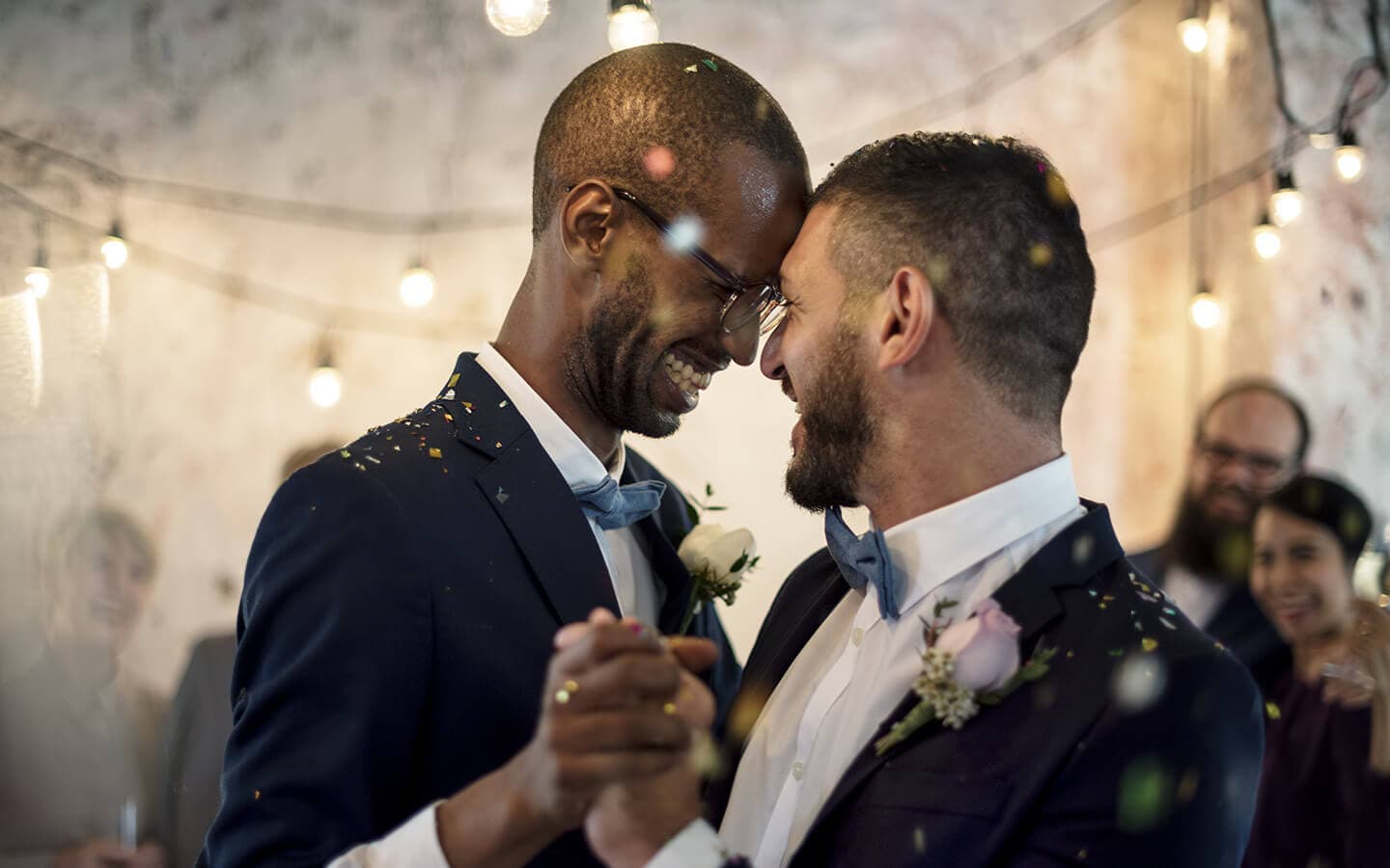 Two men share a joyful moment while dancing at their wedding, surrounded by confetti and warm lighting.