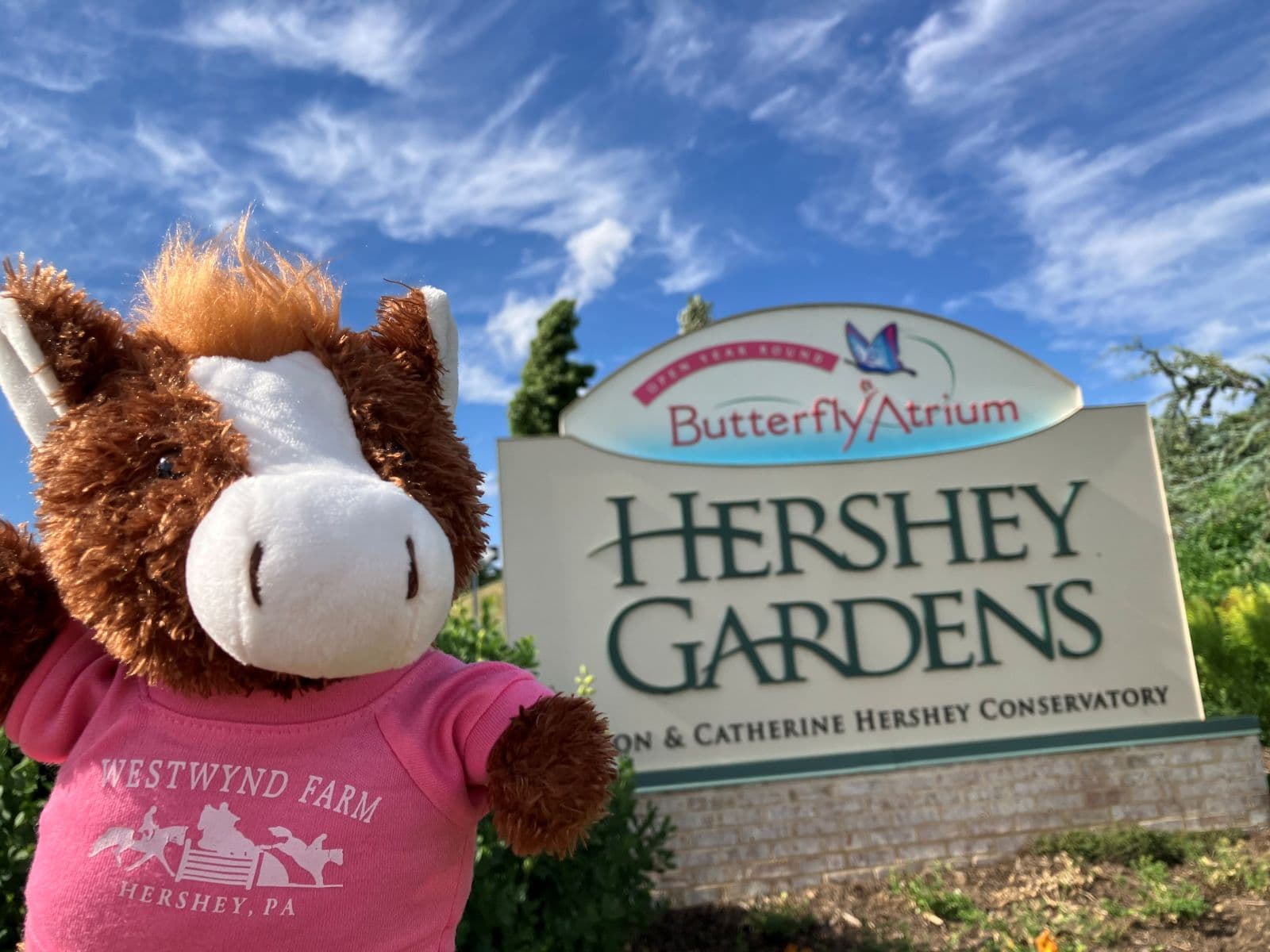 A toy horse in a pink shirt poses in front of the Hershey Gardens sign under a blue sky.