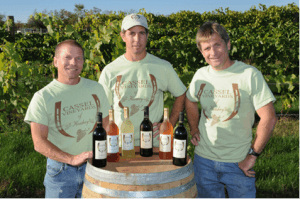 Three men in matching shirts pose with a selection of wine bottles on a barrel in front of a vineyard.