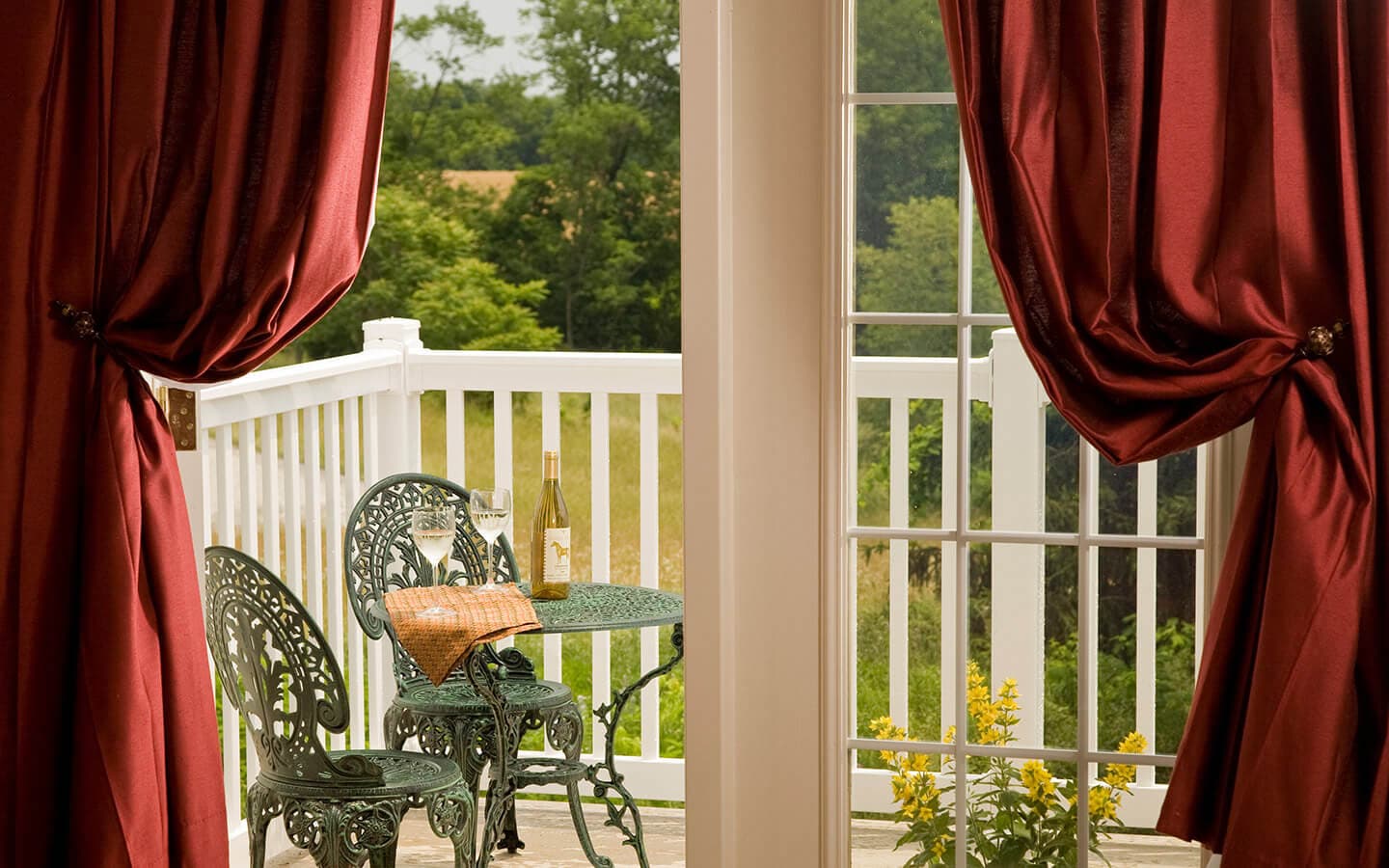 A view from inside showcasing a balcony with a small table, two chairs, and a bottle of wine, framed by red curtains.