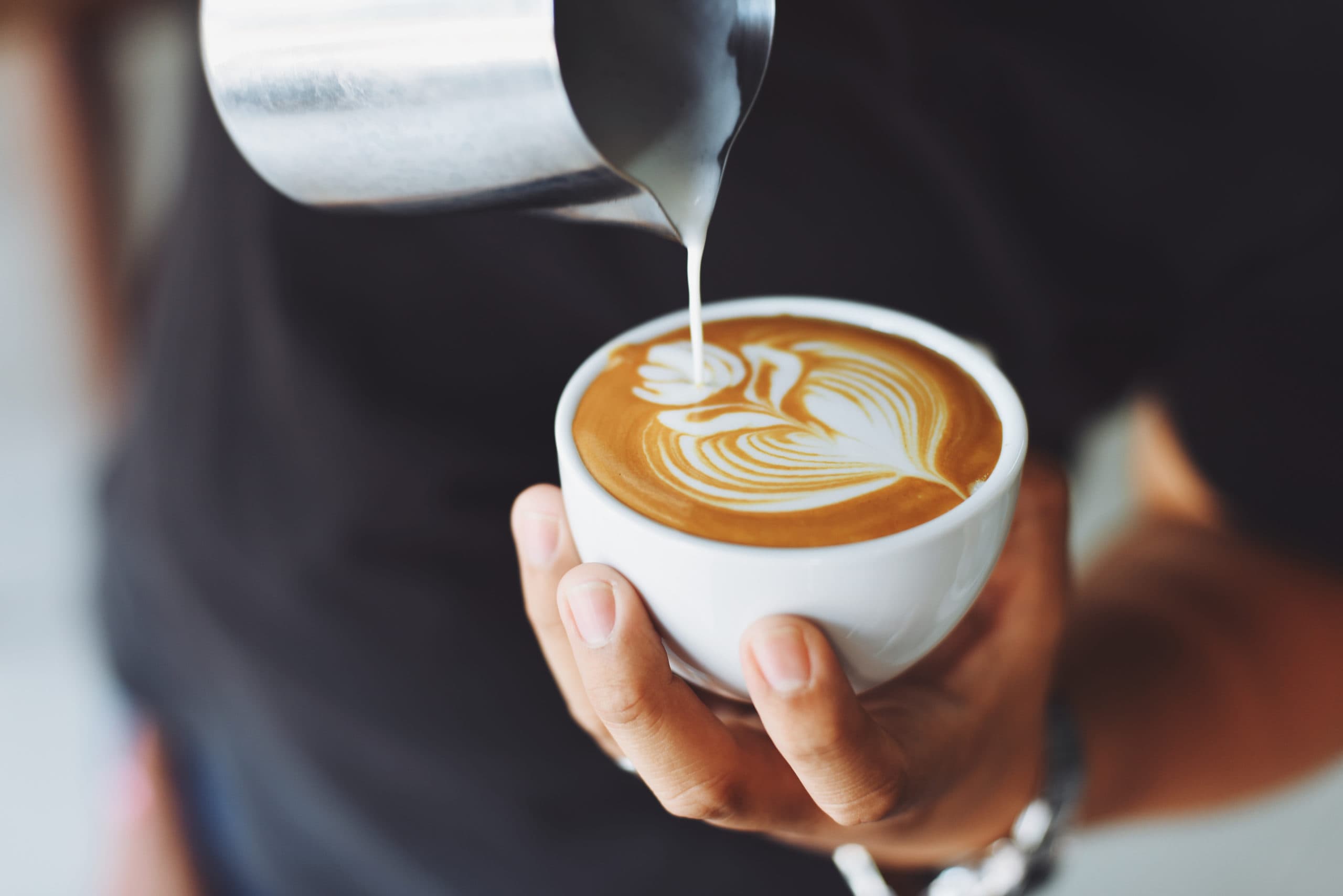 A person carefully pours milk into a cup of coffee, creating latte art.
