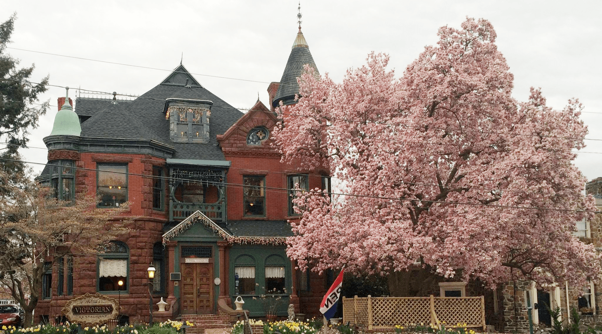 A grand Victorian house with a turret is surrounded by blooming pink flowers and greenery.