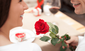 A couple shares a joyful moment over dinner, with one person holding a red rose.