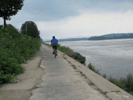A cyclist rides along a riverside path under a cloudy sky.