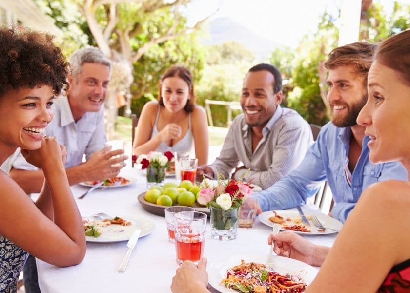 A group of six people sitting around a table enjoying a meal and drinks in a garden setting. A group of six people sitting around a table enjoying a meal and drinks in a garden setting.