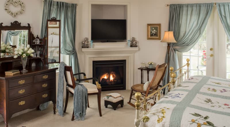 Cozy bedroom featuring a decorative quilt, fireplace, armchair, and large windows with light curtains.