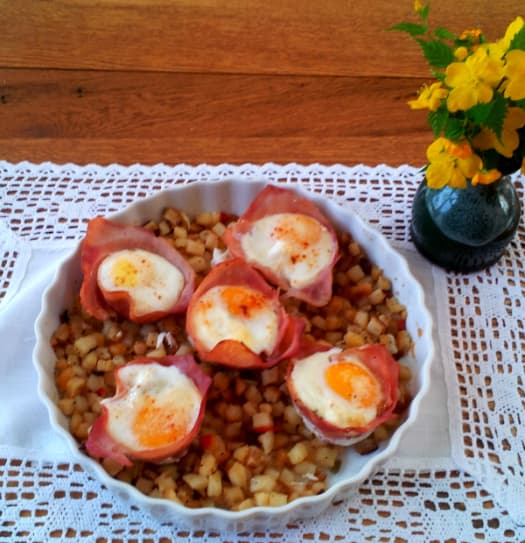 A dish of baked eggs in ham cups served over diced potatoes, with a small vase of yellow flowers nearby.