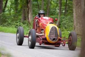 A vintage race car speeds along a winding road surrounded by lush greenery.