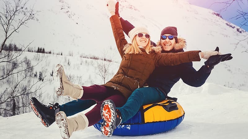 A woman and a man joyfully ride a snow tube down a snowy hill, with mountains in the background.
