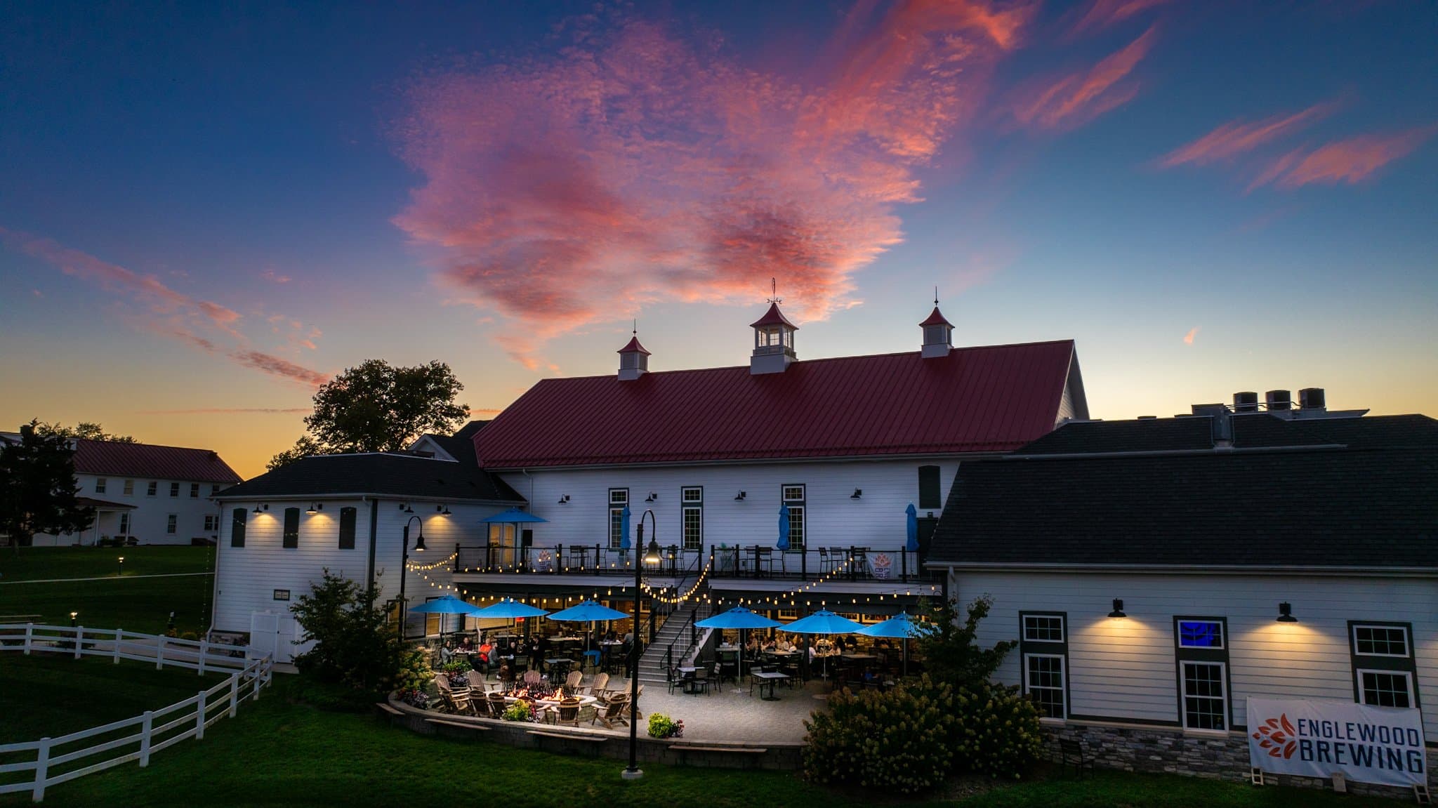 A rustic brewery with outdoor seating under blue umbrellas, set against a vibrant sunset sky.