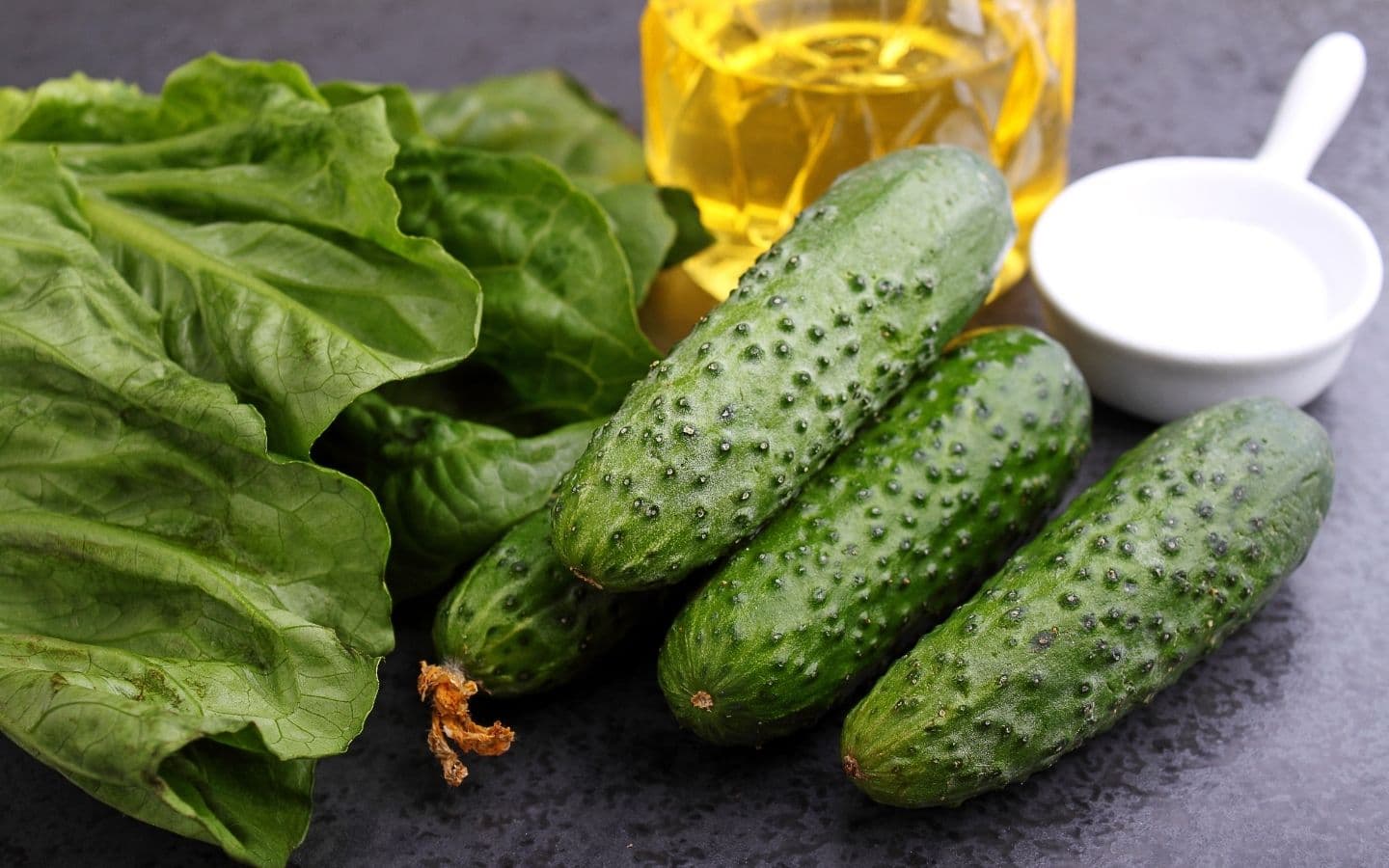 A collection of fresh cucumbers, a leaf of lettuce, a small bowl of salt, and a jar of oil. A collection of fresh cucumbers, a leaf of lettuce, a small bowl of salt, and a jar of oil.