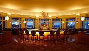 A warmly lit, circular bar area with high stools and large windows overlooking an evening landscape.