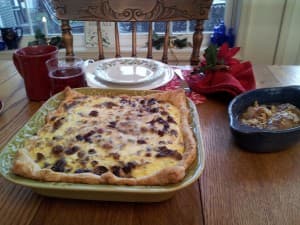 A baked dish with a golden crust and dark filling is placed on a wooden table beside mugs, plates, and a small bowl of dessert.