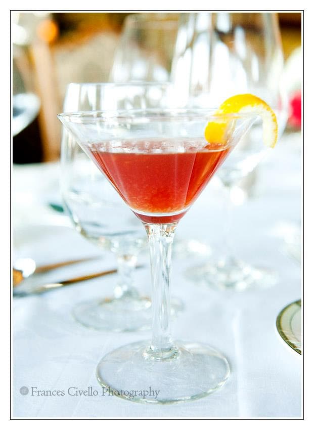 A close-up of a cocktail in a martini glass garnished with an orange twist, surrounded by other glassware on a table.