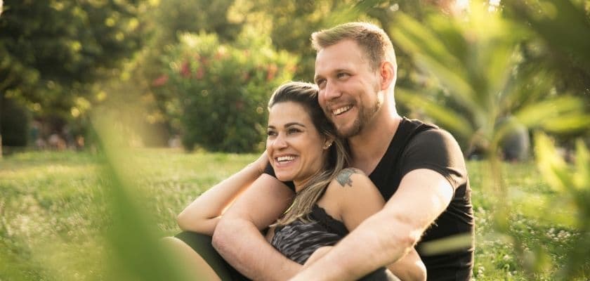A couple smiles and embraces while sitting on grass in a sunlit park.