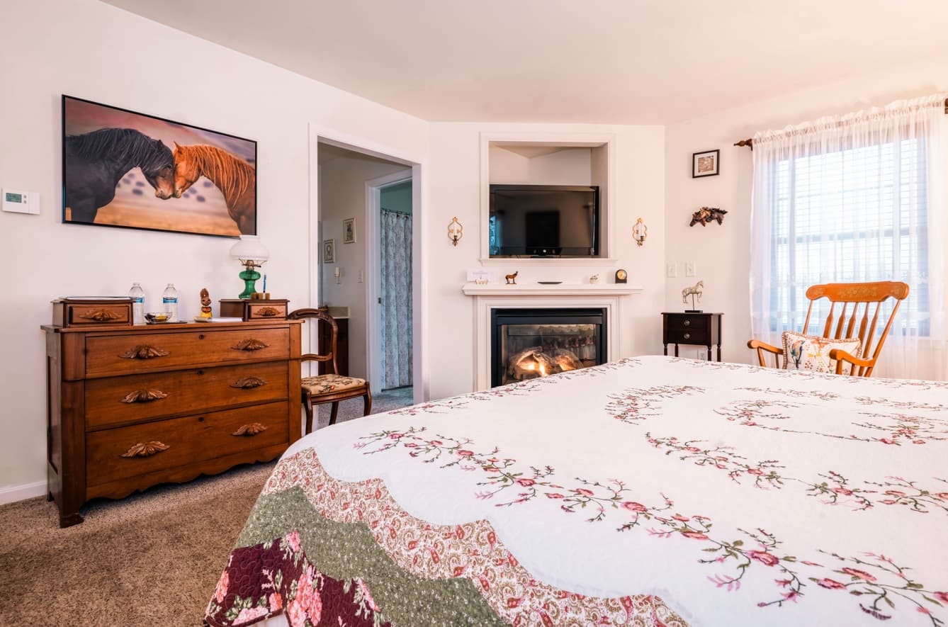 A cozy bedroom featuring a quilted bedspread, a wooden dresser, a fireplace, and horse artwork on the wall.