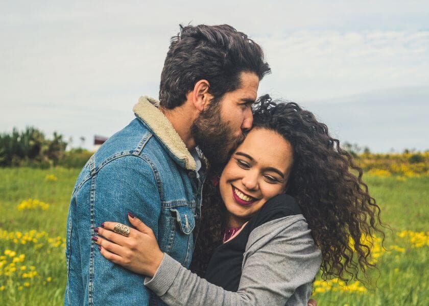 A couple embraces in a field of flowers, with the man affectionately kissing the woman's forehead. A couple embraces in a field of flowers, with the man affectionately kissing the woman's forehead.