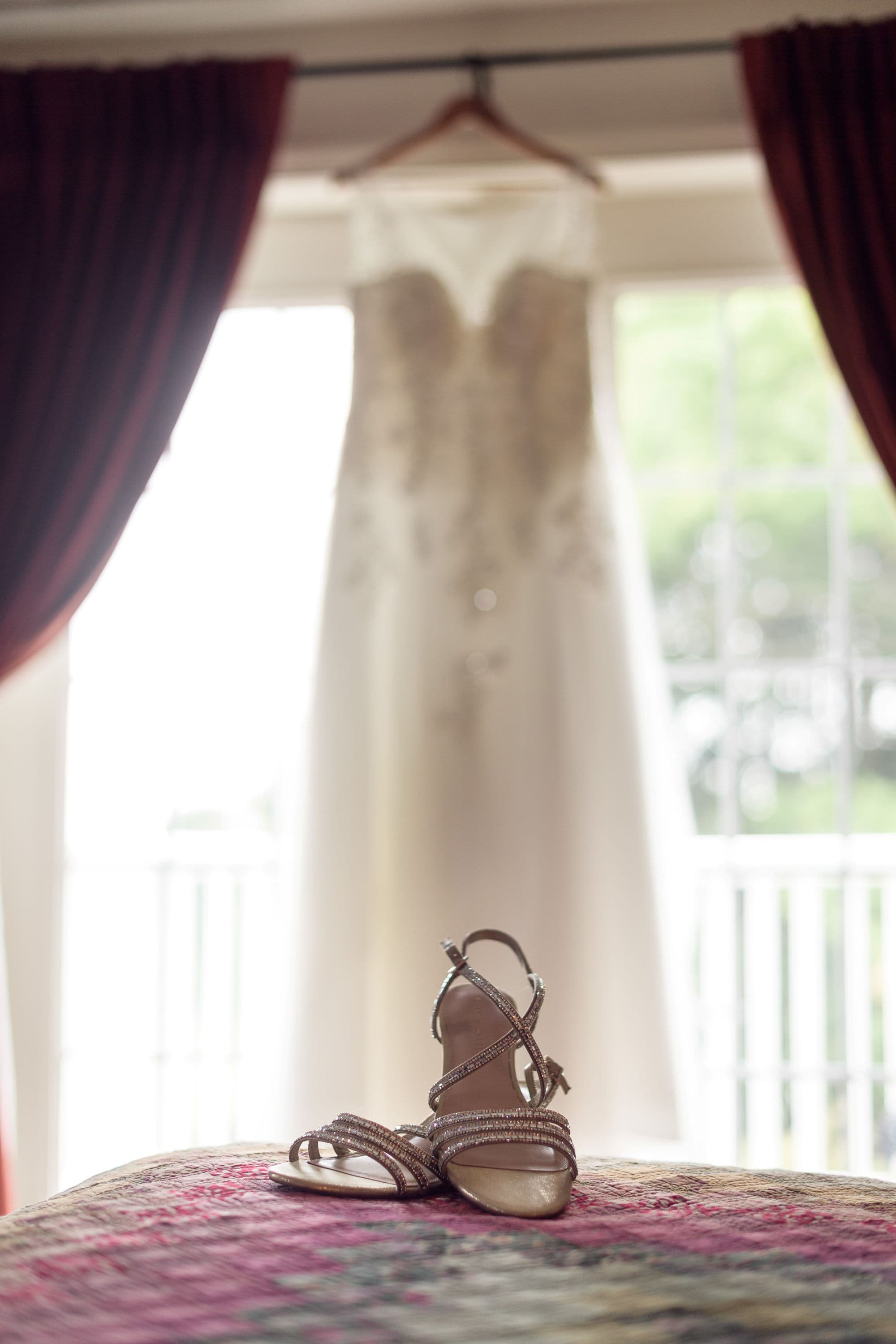 A wedding dress hangs in the background while a pair of elegant sandals rests on a colorful bedspread in the foreground.