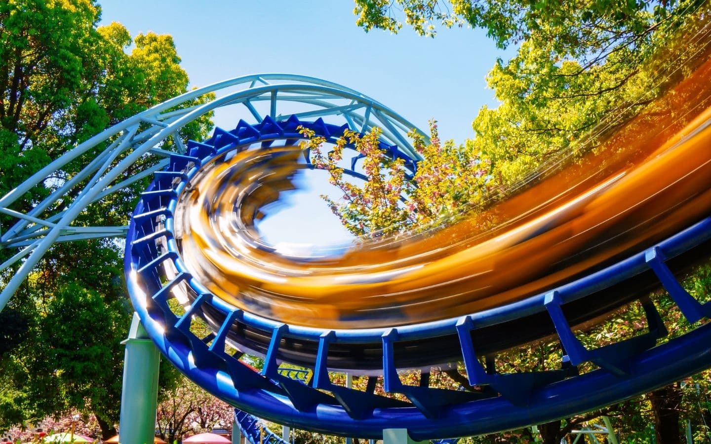 A roller coaster curves through a colorful, tree-lined landscape on a sunny day.