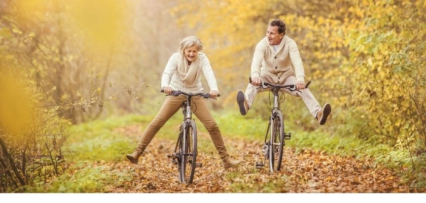 A couple joyfully rides bicycles on a leafy path in an autumn forest.