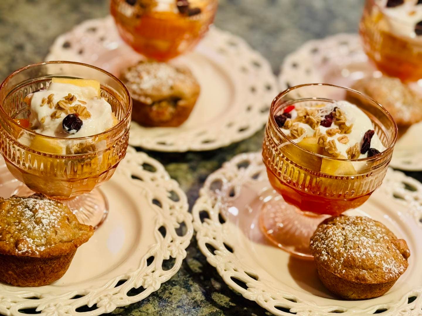 A dessert scene featuring parfaits in glass cups garnished with fruit and granola alongside muffins on decorative plates.