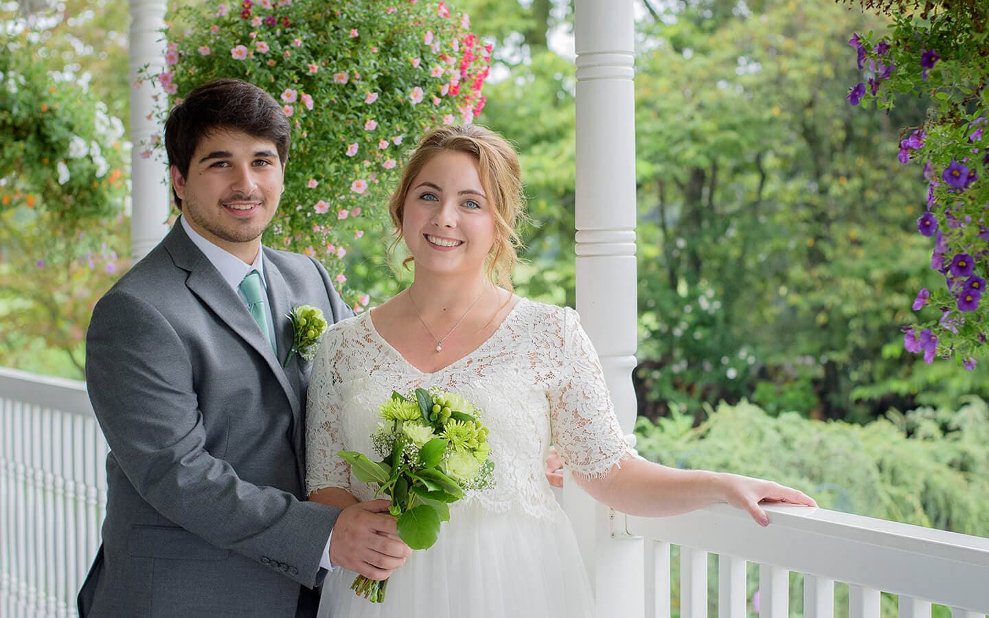 A bride and groom stand together on a porch surrounded by colorful hanging flowers.