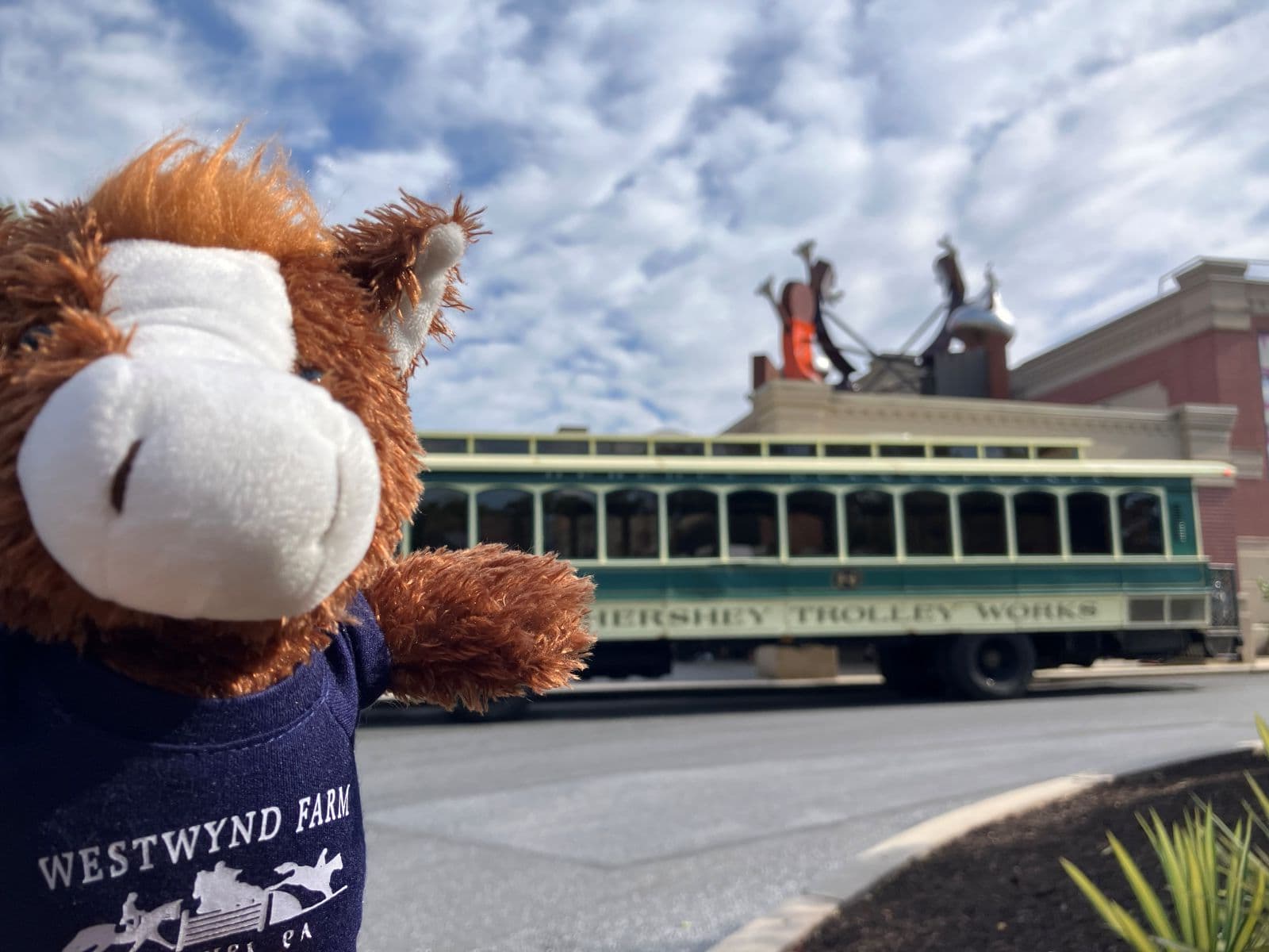 A plush horse toy in a shirt stands in front of a green trolley and a cloudy sky.