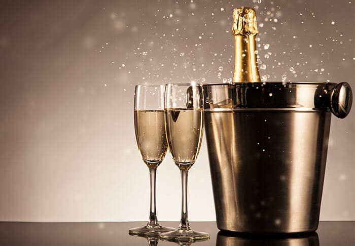 A bottle of champagne is erupting in a metal cooler next to two filled glasses.