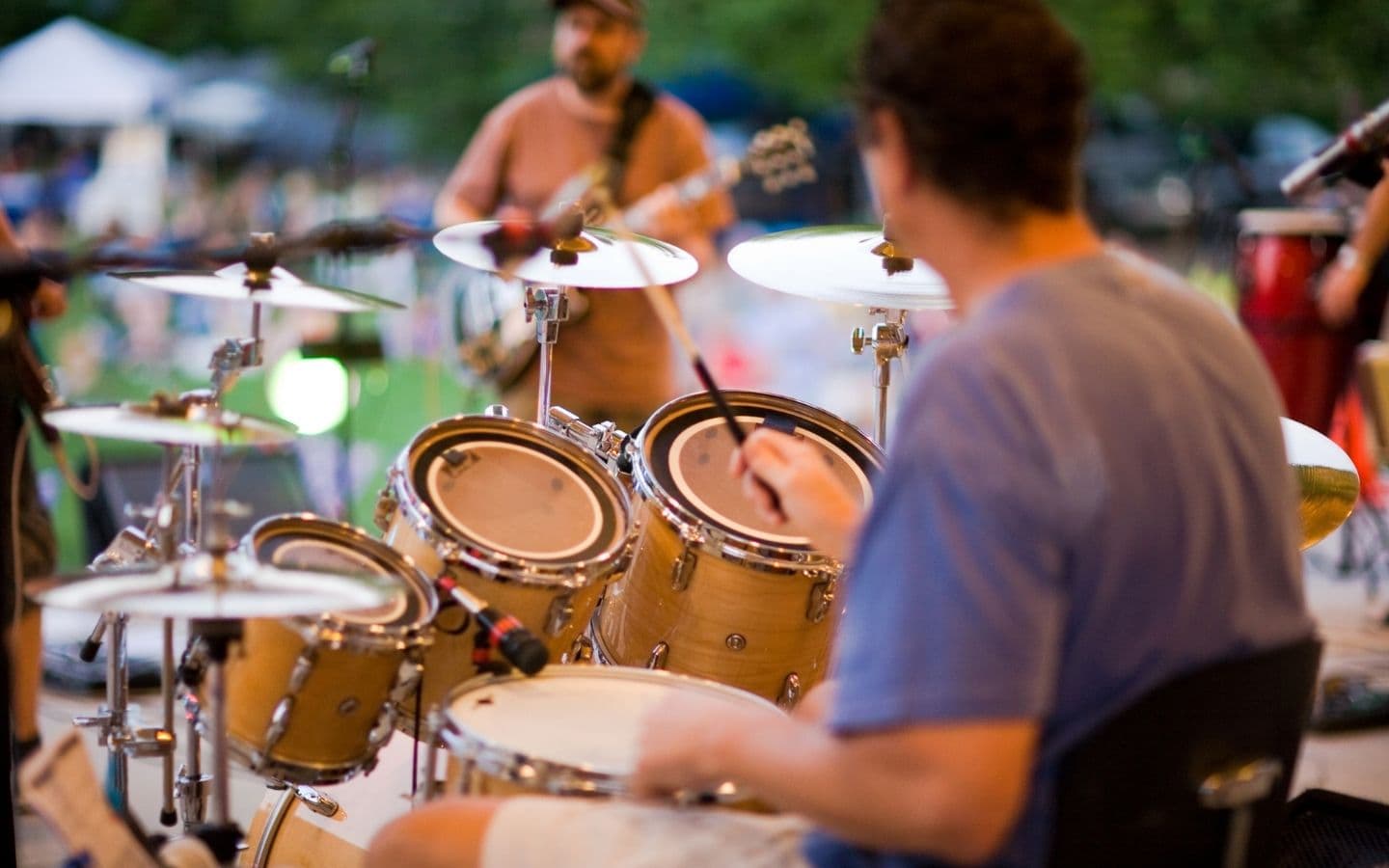 A drummer plays a wooden drum kit while a guitarist is visible in the background.