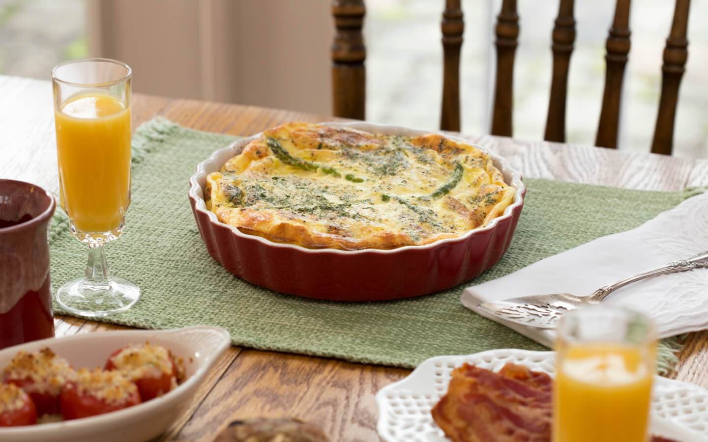 A table setting featuring a baked egg dish, a glass of orange juice, and various breakfast items.