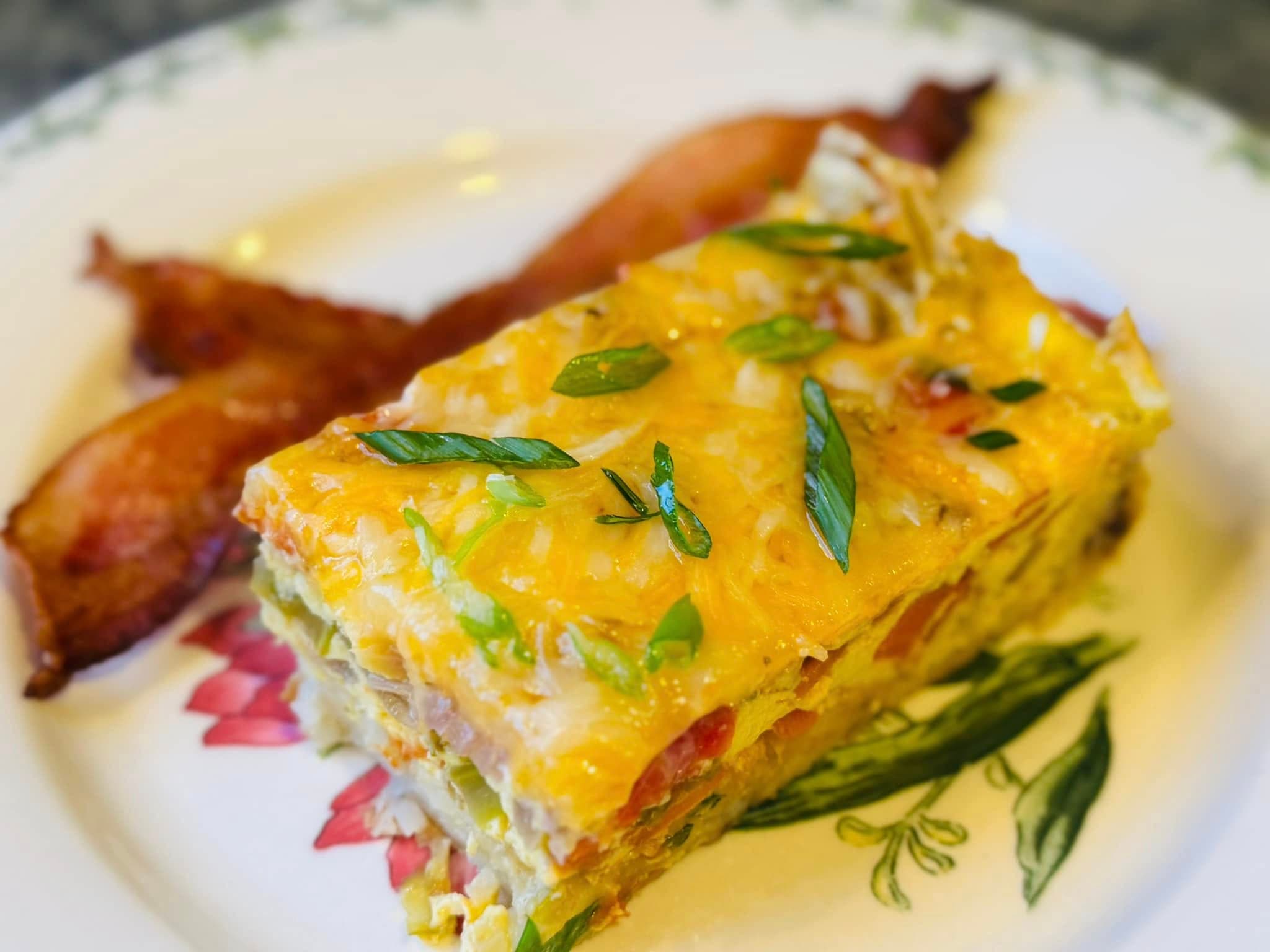 A slice of cheesy vegetable casserole with green onions served alongside crispy bacon on a decorative plate.