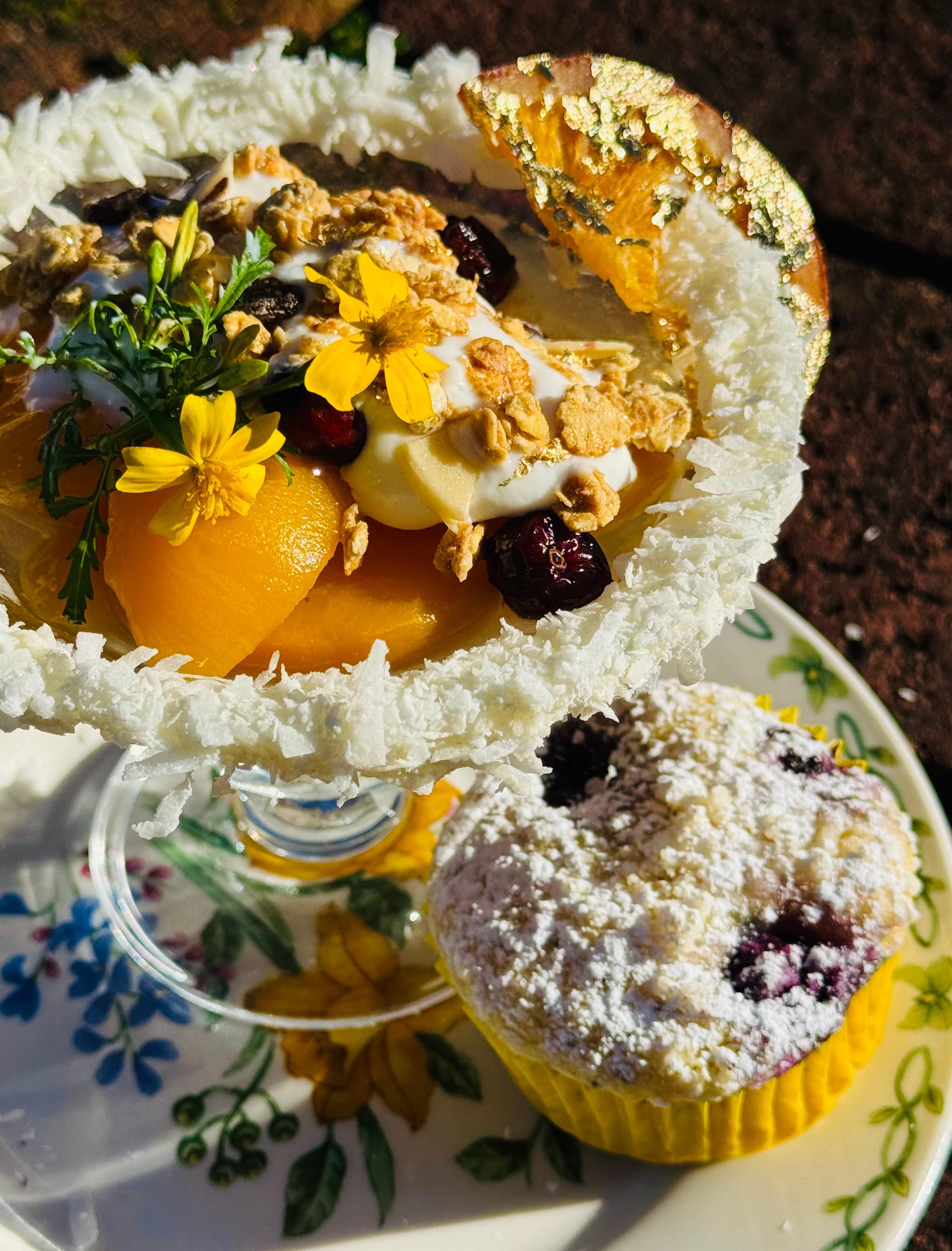 A glass dessert featuring fruit, yogurt, granola, and edible flowers, accompanied by a powdered blueberry muffin on a floral plate.