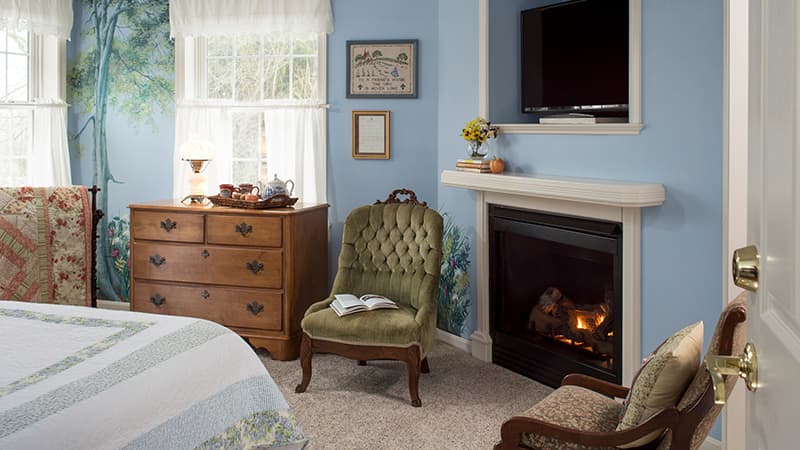 Cozy bedroom featuring a green chair, dresser, and a fireplace with a TV mounted above it.