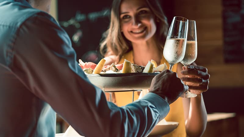 A woman smiling as she receives a plate of fruit and a glass of champagne from a person off-screen.
