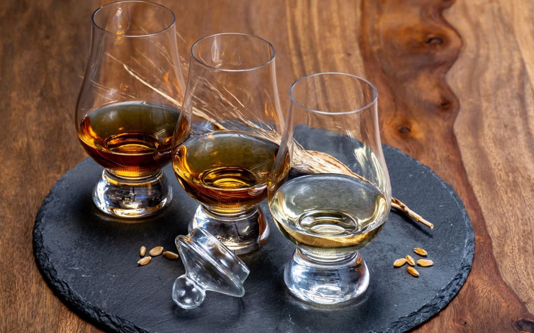 three sample glasses of distilled spirits aligned darkest to lightest left to right on a rock slab with some grains sprinkled on it on a wooden table