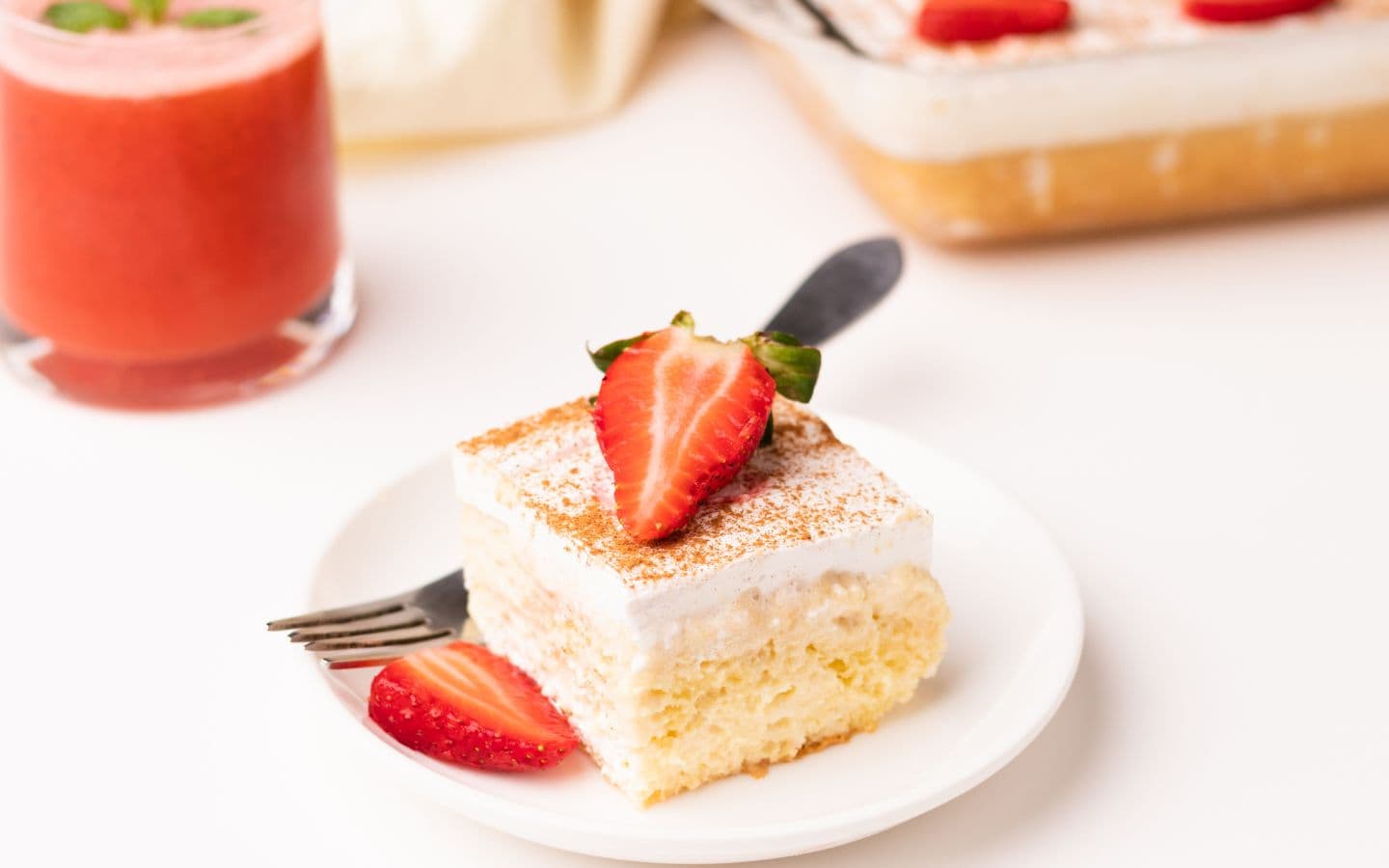 square slice of tres leches vanilla cake with sliced strawberry on top and a strawberry smoothie in the background
