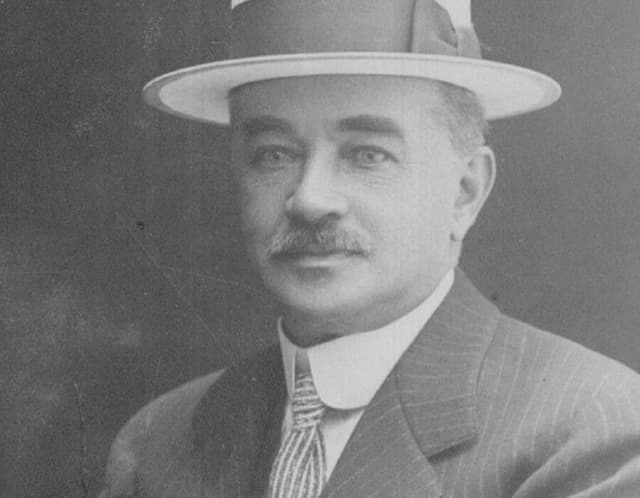 man with mustache seated with pin-striped suite and striped tie wearing hat with band