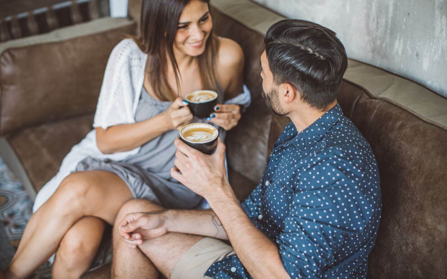 Man with brown hair in a blue button-down shirt talking to a woman with brown hair in a gray dress, both sitting on a brown leather couch drinking coffee