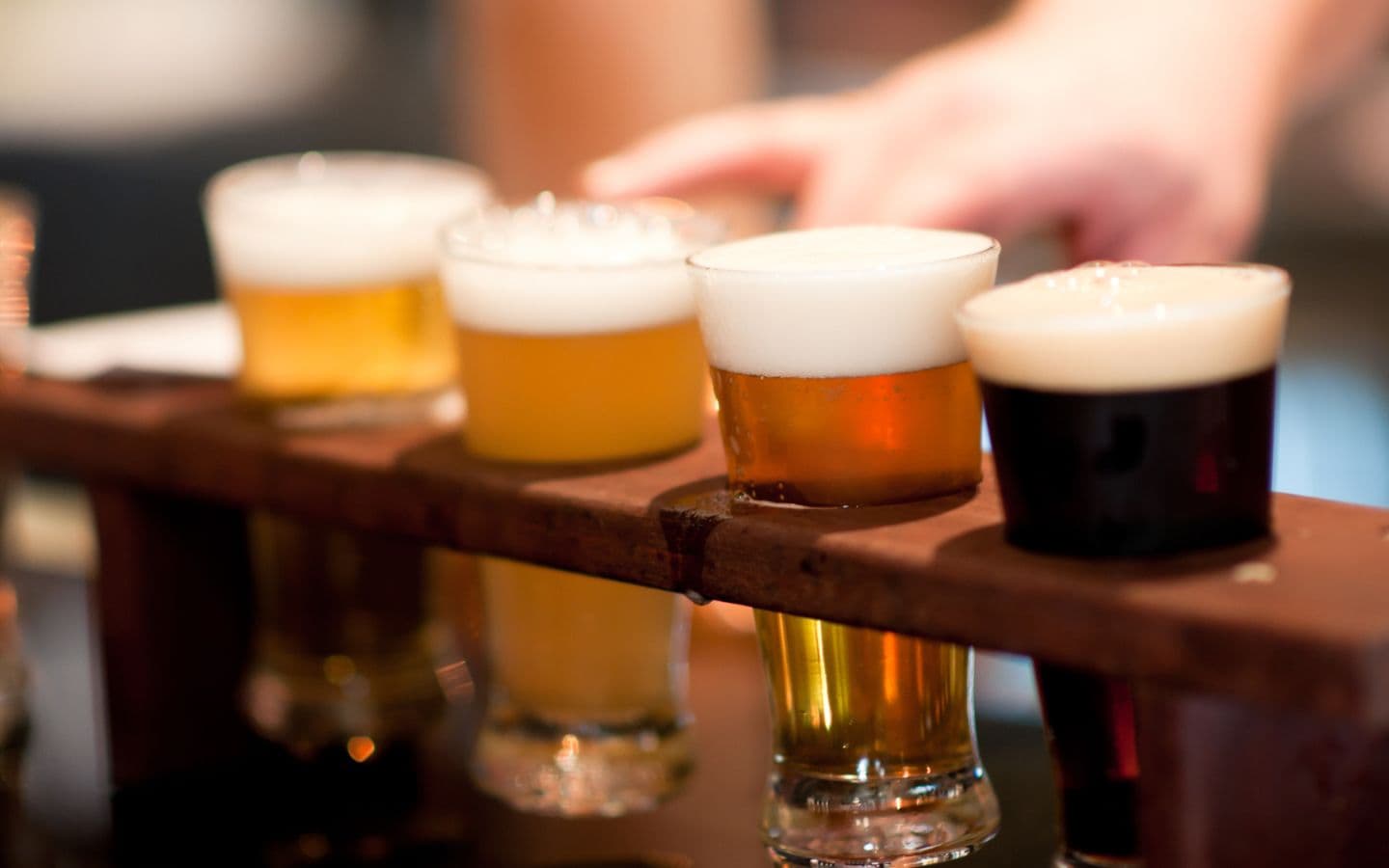 wooden beer sampler with four different beers lined up from lightest to darkest left to right and a hand reaching for the sampler in the background wooden beer sampler with four different beers lined up from lightest to darkest left to right and a hand reaching for the sampler in the background