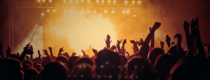 silhouette of a large crowd at a concert with hands up in front of a smokey stage with yellow lights