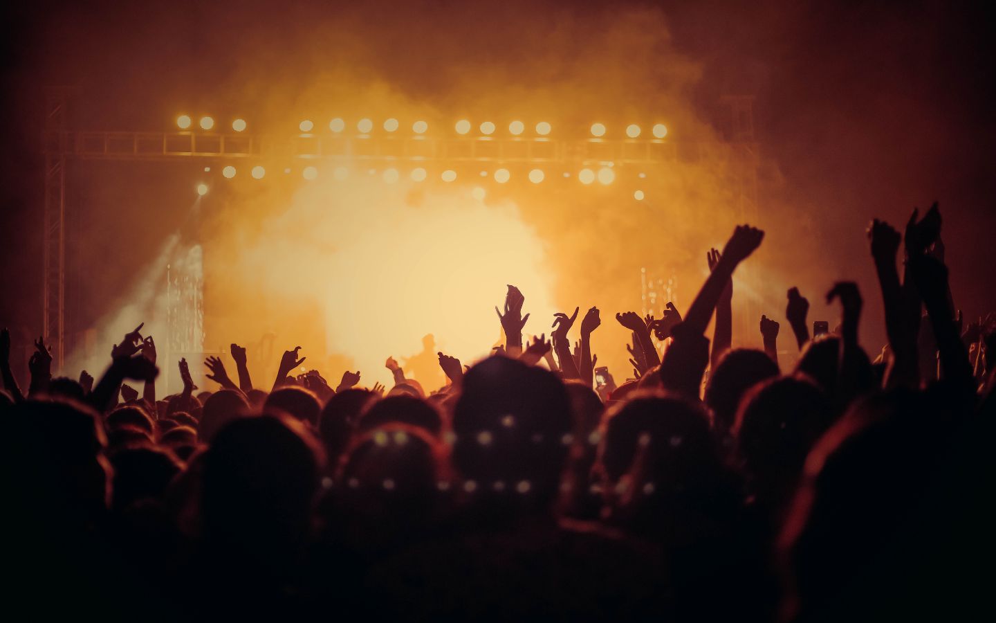 silhouette of a large crowd at a concert with hands up in front of a smokey stage with yellow lights