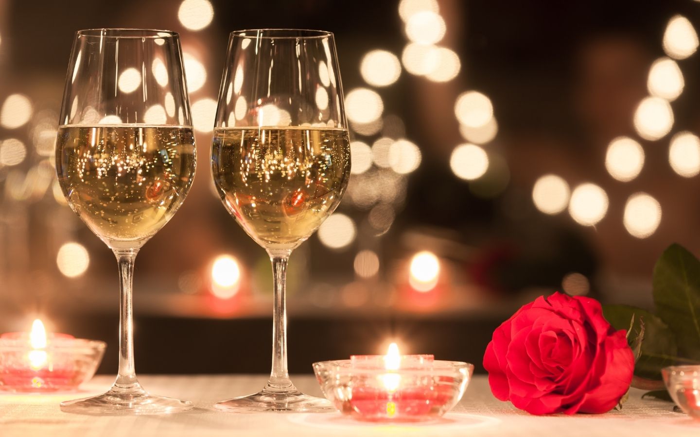 Romantic table set with wine glasses, candles, a red rose and twinkling lights in the background