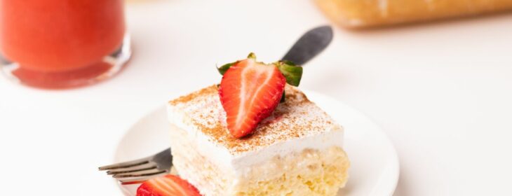 square slice of tres leches vanilla cake with sliced strawberry on top and a strawberry smoothie in the background